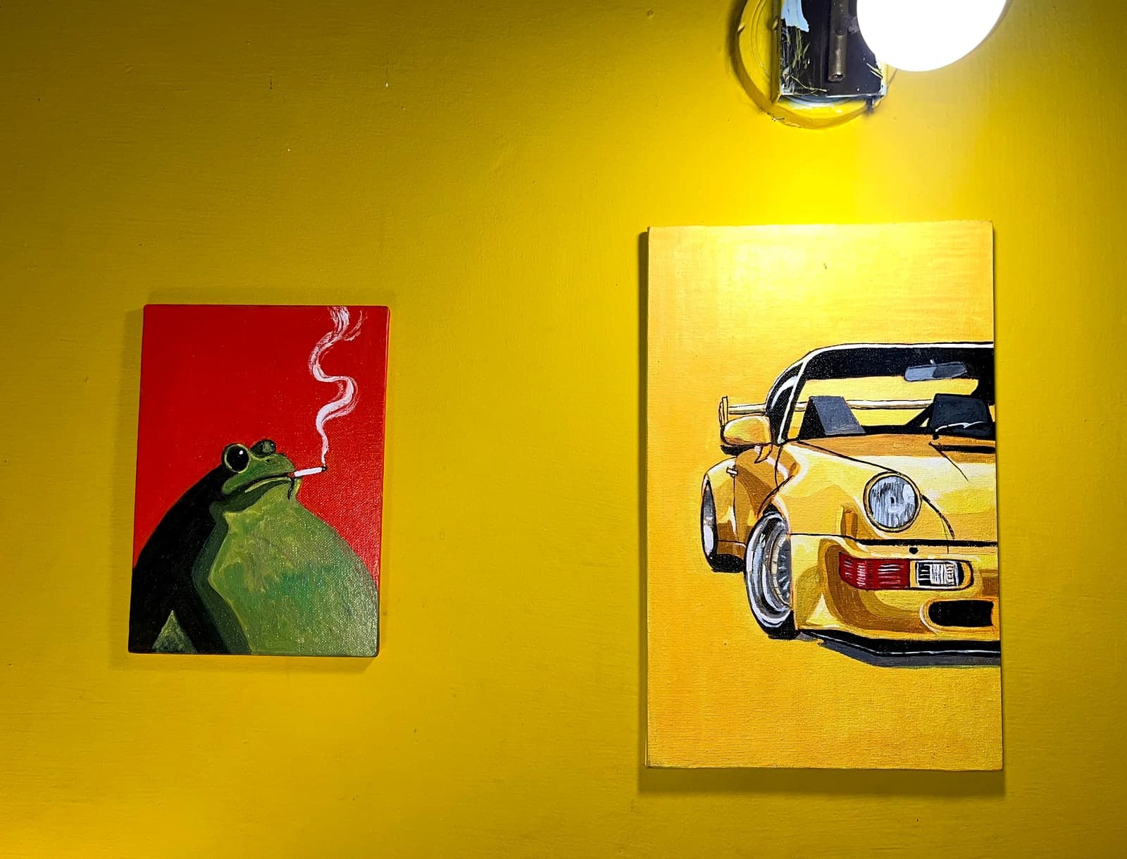 Two hand-painted acrylic canvases hanging on a bright yellow wall under a lamp: a small frog smoking a cigarette, and a larger yellow sports car
